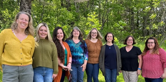 Eight women stand in a line with a forest background, smiling at the camera.