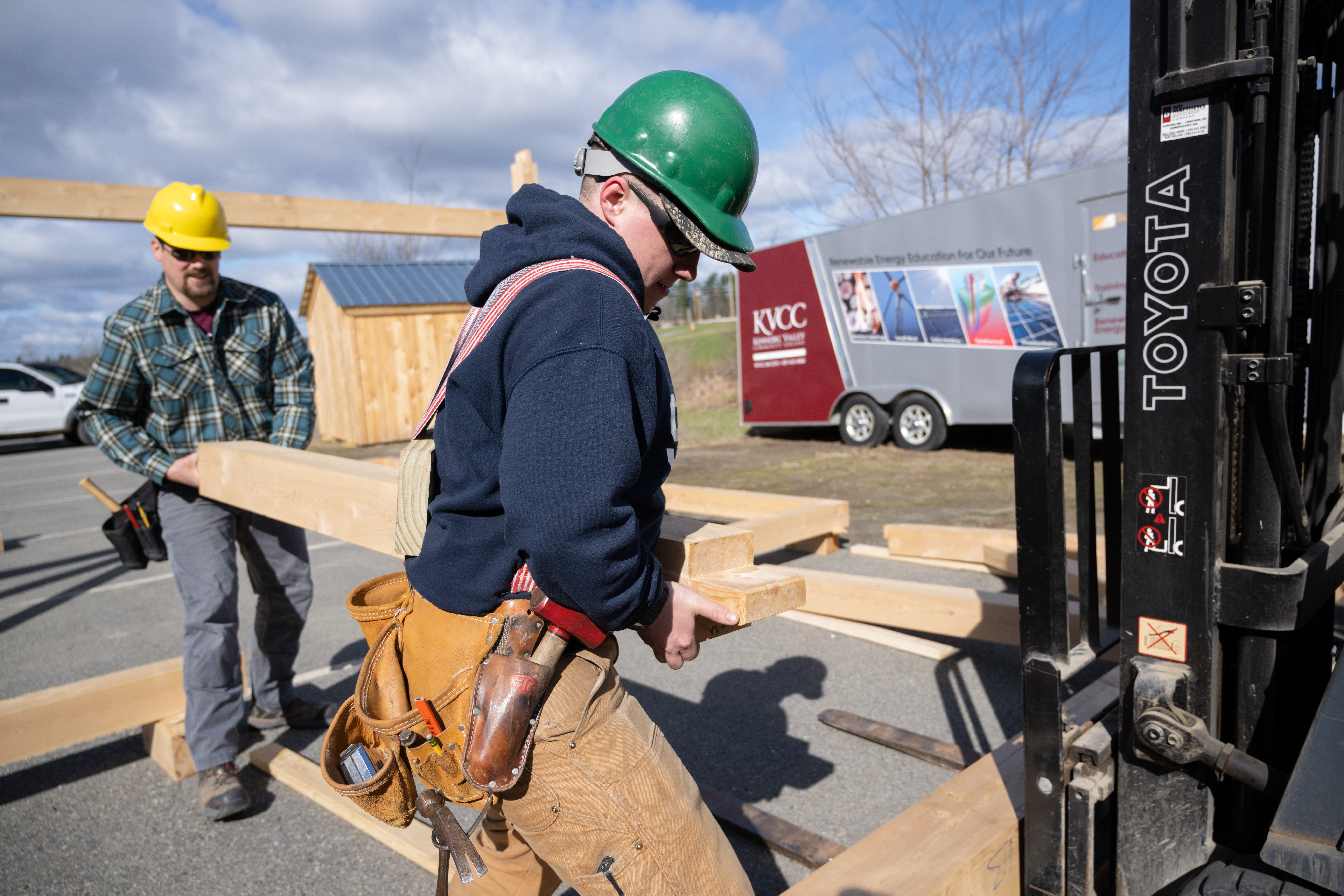 Two KVCC construction students in hard hats carry lumber outside.