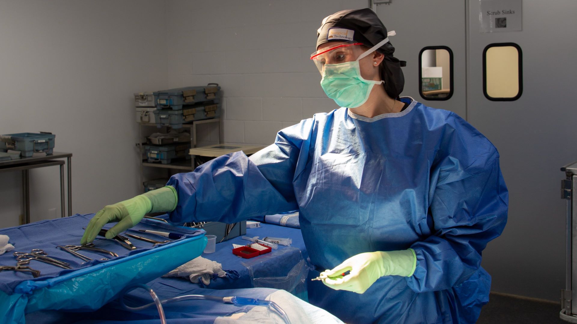Figure in blue scrubs, green mask, gloves and protective eyewear reaches for surgical tools in community college lab where students are trained as surgical technicians.