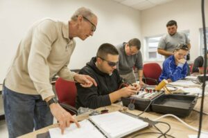 Electrician instructor stands next to four sitting students testing electrical equipment on table